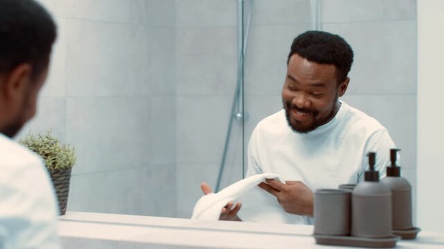 A man with dark skin is smiling and pointing at his reflection in a bathroom mirror. He is wearing a towel and appears to be enjoying his time while taking care of himself.