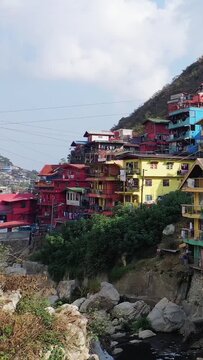 Vibrant multi-colored houses clustered on a lush hillside in Baguio, Philippines, under a bright sky. A dynamic urban landscape with natural greenery and rocky terrain.