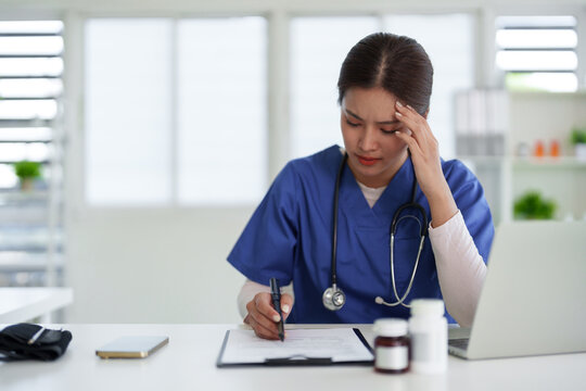 Asian adult female medical professional writing patient notes looks stressed overwhelmed modern clinic office desk healthcare workload burnout pressure