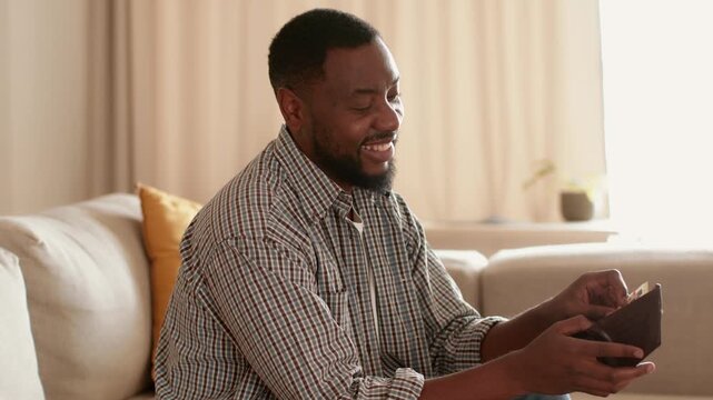 A man with dark skin sits on a comfortable sofa in a cozy living room. He smiles as he looks into his wallet, possibly checking for cash or a card. Soft light fills the space.
