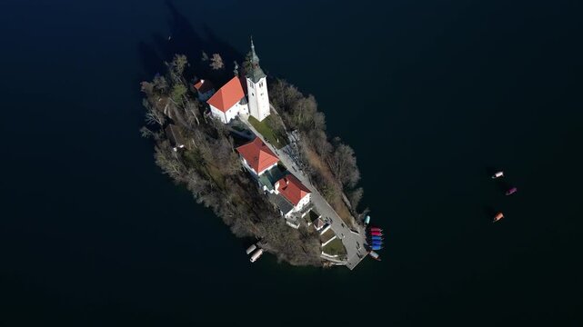 Beautiful lake Bled island in Slovenia with church and traditional pletna boats. Aerial top down view with circular motion. Calm sunny morning in snowless winter. Deep dark water in the winter.