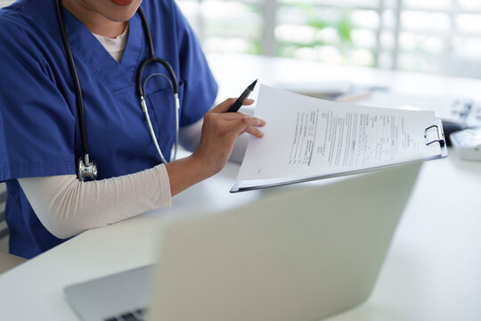 Asian adult healthcare professional reviews patient medical reports using a laptop computer at a modern clinic office desk for diligent recordkeeping data entry