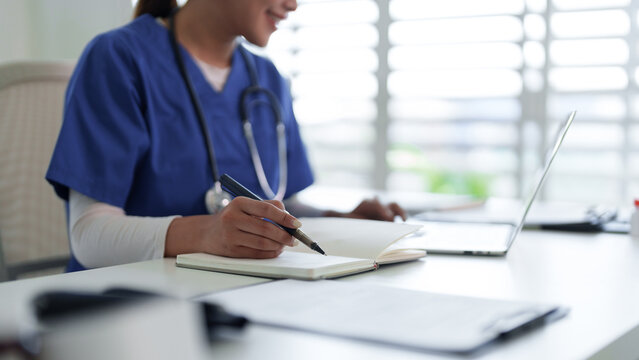 Asian medical professional adult woman writing notes notebook using laptop computer studying healthcare clinic office workspace