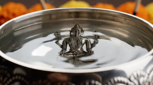 Mahavir Jayanti theme, close-up of a drop of water falling into a silver bowl during Abhisheka (ritual bath). The focus is on the ripples and the reflection of a Mahavira statue in the water.