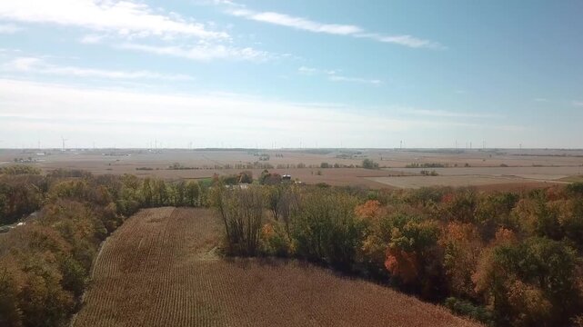 Aerial view of a vast agricultural landscape with fields of harvested crops, vibrant autumn trees, and numerous wind turbines on the horizon under a clear blue sky