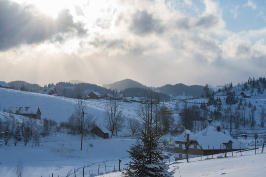Snowy rural mountain village landscape with traditional houses, pine trees and cloudy winter sky