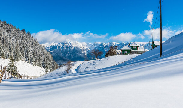 Snowy mountain village landscape with wooden houses, pine forest, and distant peaks under clear blue sky in winter