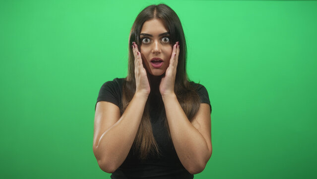 Woman covers mouth with hands, wide eyes and burgundy painted nails facing camera against green studio backdrop; surprise curiosity.