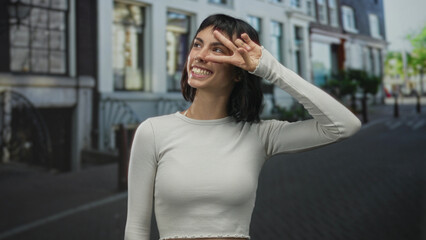 Fototapeta premium Woman shows v sign with hand to face while smiling on a sunlit cobblestone street; carefree happiness.