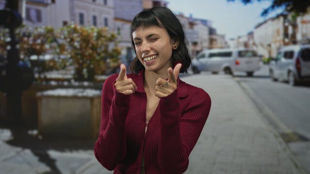 Young hispanic woman with smiling face pointing fingers at camera on urban street; playful confidence.