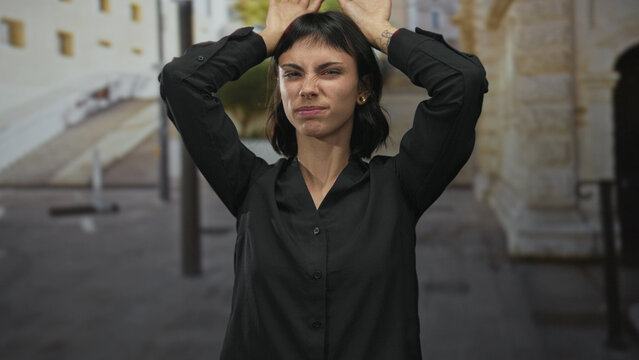 Young hispanic woman making rabbit ears gesture with bare hands atop head on bustling street; playfulness.
