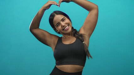 Fototapeta premium Young hispanic woman with bare arms and hands forms heart with fingers above her head while smiling, wearing a black sports bra in a studio setting; joy wellness positivity.