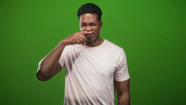 Man pinches nose in studio with green backdrop and pink tshirt, grimacing at a foul smell; disgust discomfort.