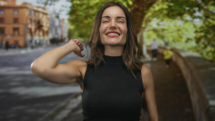 Naklejka premium Woman pointing finger at camera with raised arm on a sunlit city street flanked by trees and buildings; confidence.