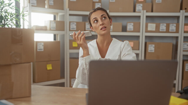 Woman holding phone and speaking into it above a parcel at a building desk surrounded by stacked boxes and shelves; small business stress.