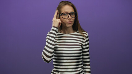Naklejka premium Woman pointing finger to temple and pointing at camera in studio with purple backdrop wearing striped top and black glasses; thinking focus.