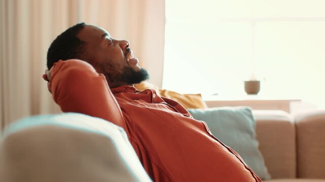 A man with dark skin sits comfortably on a couch in a well-lit living room. He leans back with a smile, enjoying a moment of relaxation and comfort.