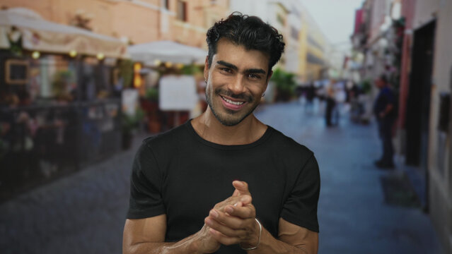 Man wearing black shirt and silver bracelet claps hands while smiling on street during a sunny afternoon; celebration.