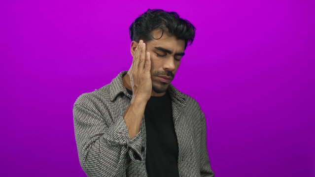 Young hispanic man grimacing and pressing hand to cheek for toothache in purple studio setting; discomfort.