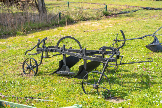 Vintage plow rests in sunlit field. Rusted metal frames bear farm history. Green grass stretches under bright sky. No people, only stillness and silence. Perfect for rustic, agricultural