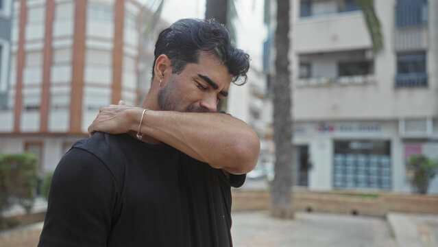 Man touches neck and winces on sunlit city street by building facade wearing a black shirt; discomfort.