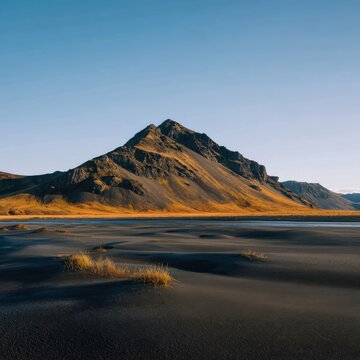 Golden light on black sandy beach with grassy tufts and sloping mountain under a clear pale sky in a tranquil coastal scene