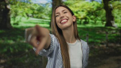 Naklejka premium Young woman in white shirt smiles while making heart gesture with her hands in green forest; affection love.