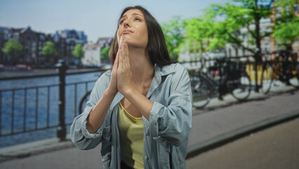 Fototapeta premium Young hispanic woman with hands clasped in prayer and closed eyes on amsterdam street by canal railing, wearing denim shirt; hope plea.