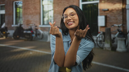 Fototapeta premium Woman points fingers at cobblestone street, makes finger heart and beckoning gestures while smiling, wearing glasses and denim shirt; playful charm.