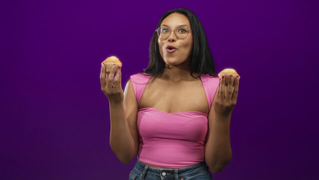 Woman holding muffins in hands with mouth open surprised expression in studio wearing pink top and jeans; playful delight.