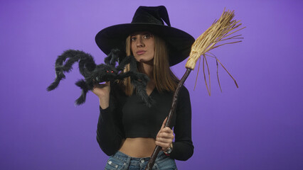 Fototapeta premium Woman in witch hat holds large fake spider puppet and straw broom while exposing midriff in studio purple set; playful charm.
