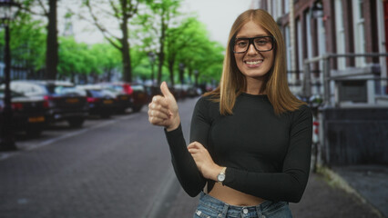 Fototapeta premium Woman redhead smiling shows thumbs up with her hand and crossed arms visible next to parked cars on a city street; confidence.