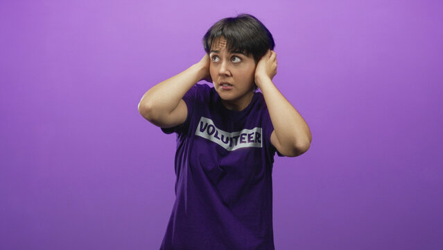 Woman volunteer in purple shirt covers ears with hands and grimaces in a studio while flinching from noise and looking distressed; anxiety overload.