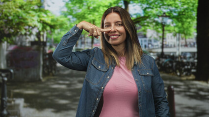 Fototapeta premium Smiling woman points finger to cheek on busy city street wearing denim jacket and pink shirt while looking at camera; playful joy.