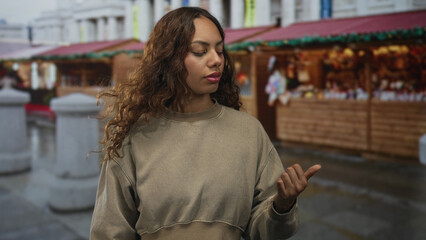 Fototapeta premium Woman with palms up and closed eyes standing on street in front of wooden souvenir stalls and stone bollards, wearing beige sweatshirt; calm reflection.