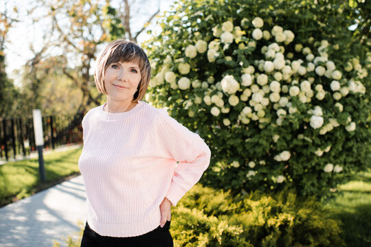 Blonde senior woman 55-60 year old posing with blooming bushes with white flowers in park outdoors. Mature wellbeing lifestyle. Springtime.