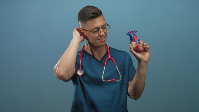 Man cardiologist in studio holding anatomical heart model, hand to neck and stethoscope visible; playful curiosity.