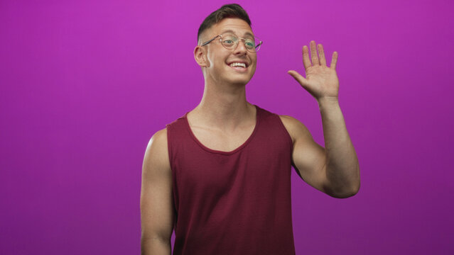 Young man in maroon tanktop and glasses points finger upward, bare arm visible, smiling in studio purple backdrop; greeting cheerful.