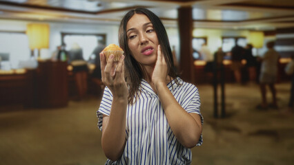 Fototapeta premium Young hispanic brunette woman holding muffin, hand on cheek, wearing striped shirt in hotel building; toothache discomfort.