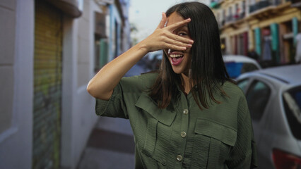 Fototapeta premium Hispanic woman covers eyes with hand on a city street while smiling beside parked cars and colorful facades; playful surprise.