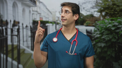 Obraz premium Man doctor with stethoscope points finger upward on street with townhouse railings; confidence care.