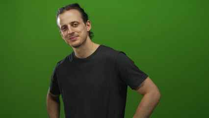 Fototapeta premium Man with hands on hips and visible bare forearms in studio with green backdrop, slight smile and relaxed posture; confident approachable.