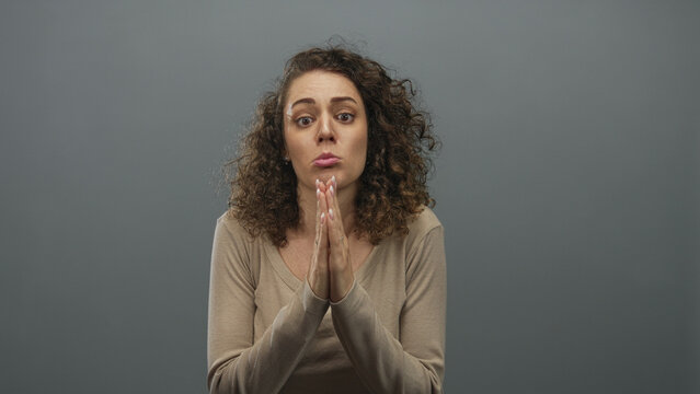 Young hispanic woman pressing hands together in pleading gesture wearing beige top in gray studio; remorse apology.