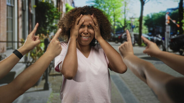 Woman with hands on head and multiple fingers pointing at her on a busy city street with parked cars; isolation anxiety.