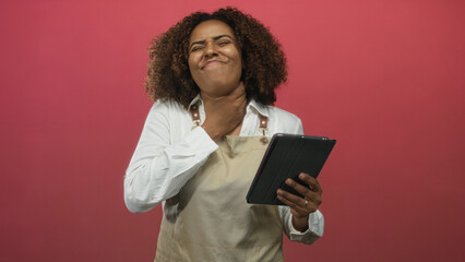 Woman baker in apron clutching neck and holding tablet in pink studio, eyes closed and face grimacing  pain stress. © Krakenimages.com