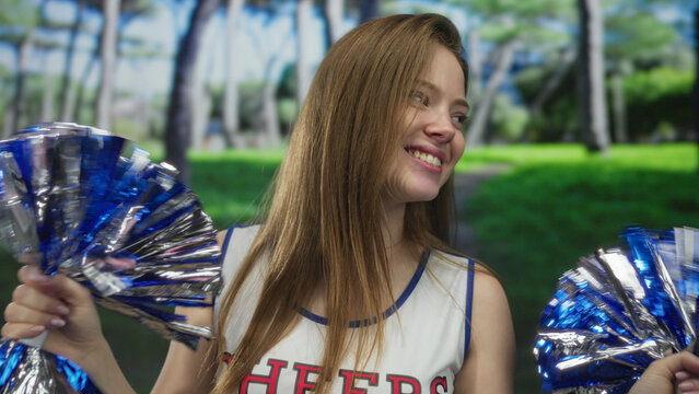 Young caucasian woman shakes blue and silver pom poms energetically while smiling in green park; team spirit joy.