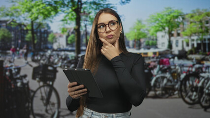 Fototapeta premium Woman wearing glasses and black top taps tablet with finger while holding device against a studio backdrop of parked bicycles; thoughtful.