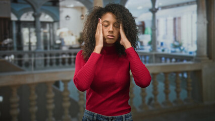 Fototapeta premium Teen woman with curly hair pressing hands to temples in ornate building wearing red sweater; stress headache.