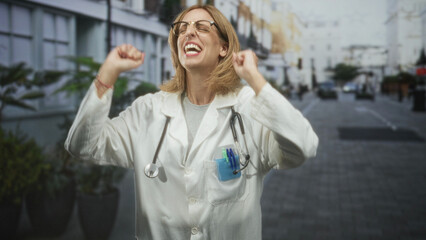 Fototapeta premium Woman doctor in white coat with stethoscope raises hands and smiles on a city street; joy relief success.
