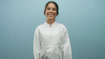 Fototapeta premium Woman chef in white double breasted coat smiles softly at camera against a pale blue studio wall; friendly confidence.